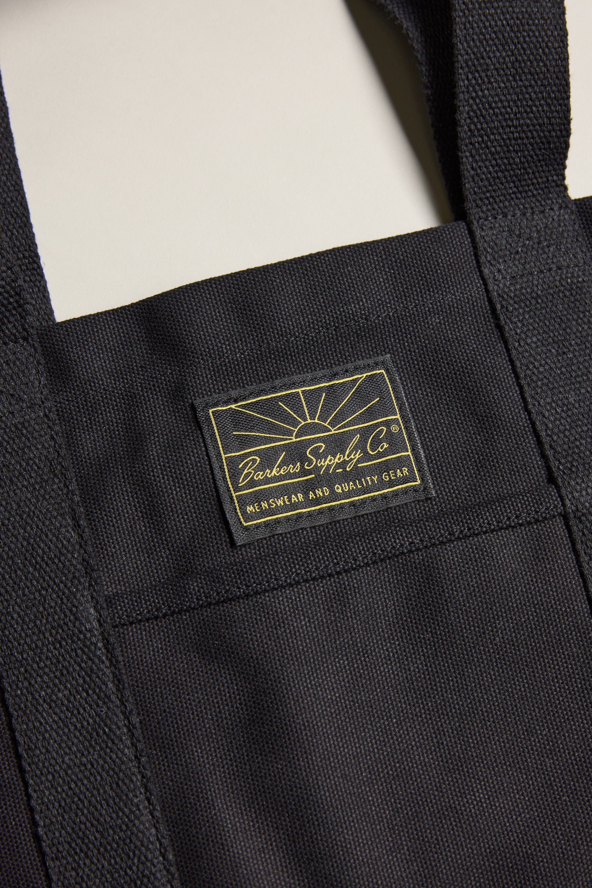 Close-up of the Barkers Supply Co. Canvas Tote Bag by Barkers, made from durable black canvas with sturdy handles, visible stitching, and a sewn label featuring yellow text and a sunrise design.