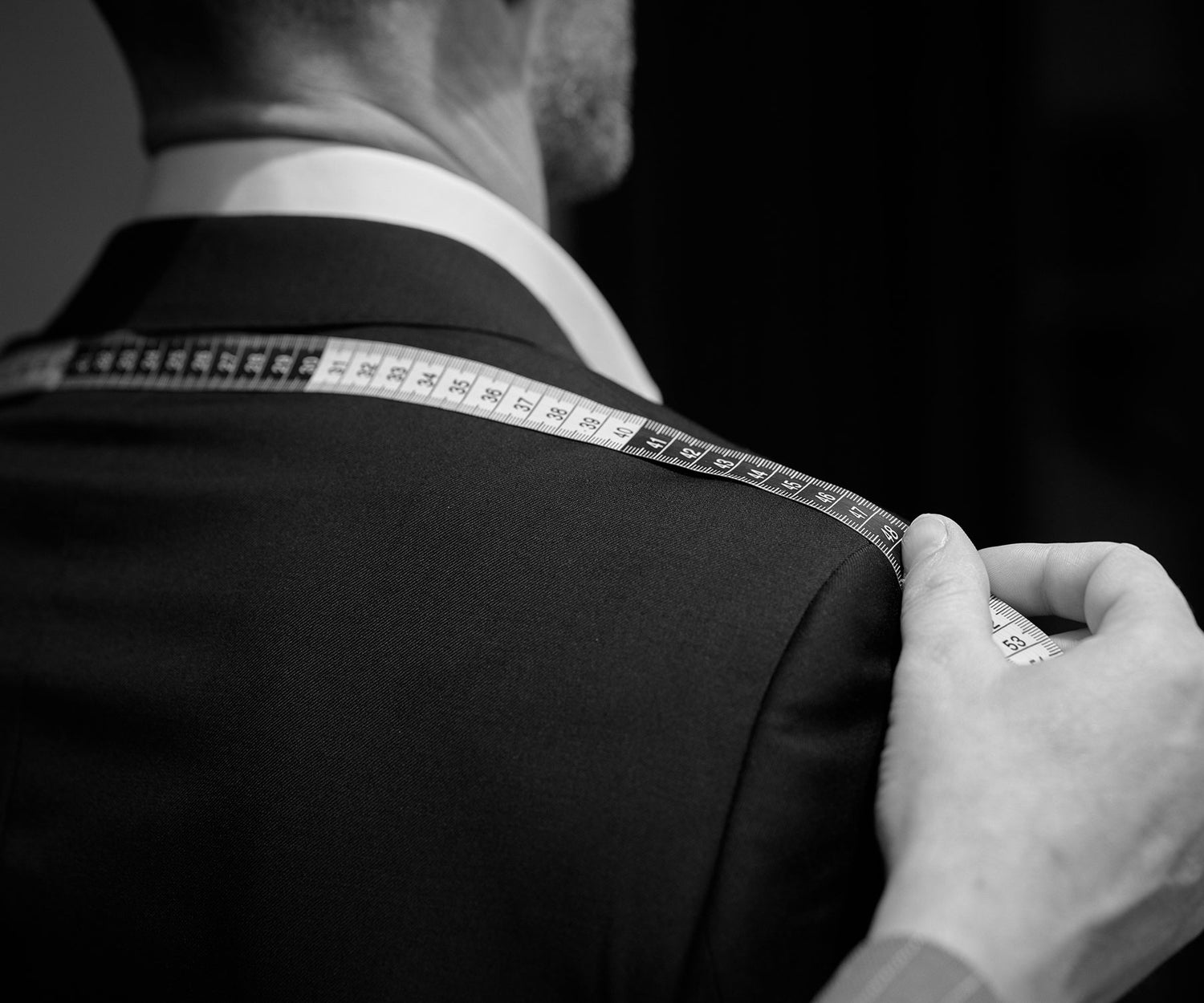 A tailor measures a mans shoulder with a measuring tape, preparing to fit or alter his suit. The image is in black and white and focuses on the back of the mans suit.