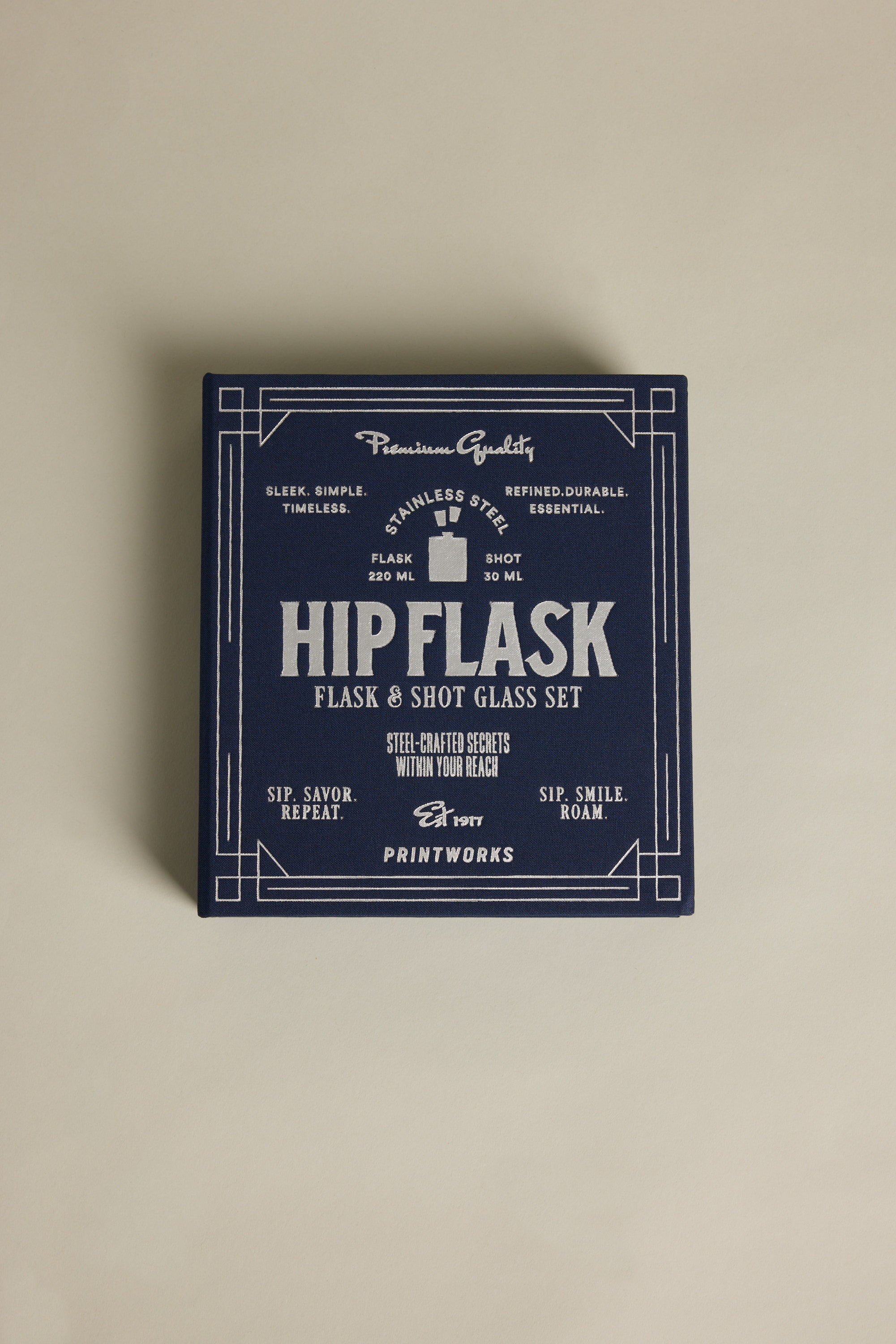 A navy blue box with "The Essentials Hip Flask" in white vintage-style text by Barkers highlights a stainless steel flask and shot glass set, showcasing quality, craftsmanship, and timeless appeal.