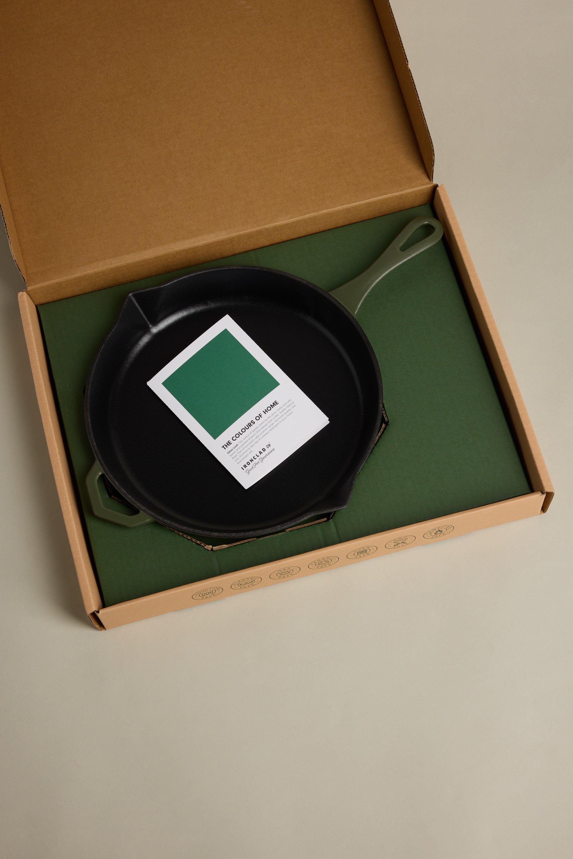 An IronClad Satin Enamel Cast Iron - 28cm Legacy Pan satin with a green handle rests in an open box on green packaging, with an informational card placed inside the induction-ready cookware.