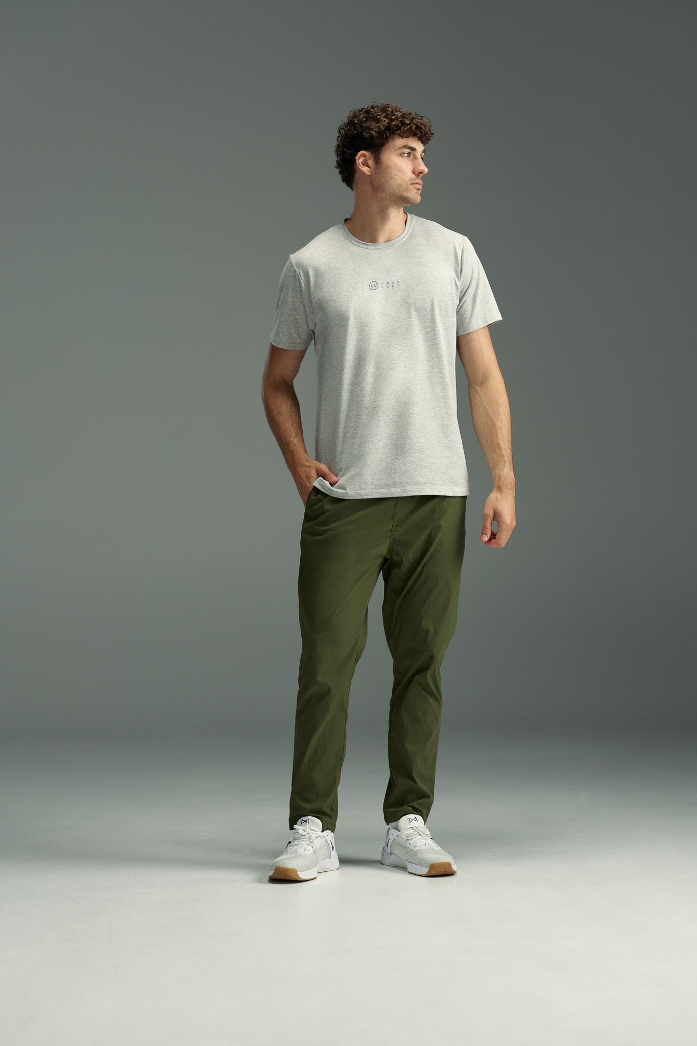A man with curly hair stands on a gray background, wearing a light gray t-shirt, white sneakers, and olive green WearEver Transit Pants from WearEver. He has one hand in his pocket and looks off to the side.