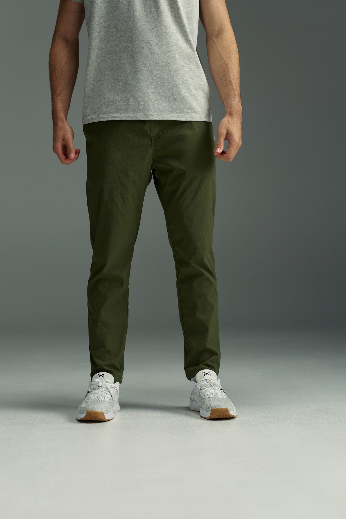 A person stands against a gray background, wearing the WearEver Transit Pant by WearEver in olive green with a light gray tee and white sneakers. The photo, cropped at the shoulders, highlights the lightweight pants and relaxed style.