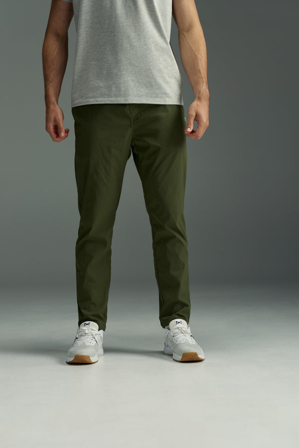 A person stands against a gray background, wearing the WearEver Transit Pant by WearEver in olive green with a light gray tee and white sneakers. The photo, cropped at the shoulders, highlights the lightweight pants and relaxed style.