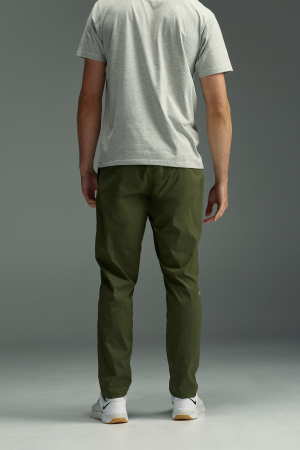 A person stands facing away, wearing a light gray T-shirt, WearEver Transit Pant in olive green, and white sneakers with black accents, against a plain gray background.