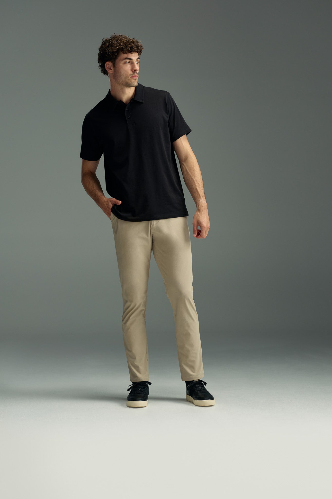 A man with curly hair in a WearEver Outbound Polo, beige pants, and black sneakers stands with one hand in his pocket, gazing to the side against a plain gray background.