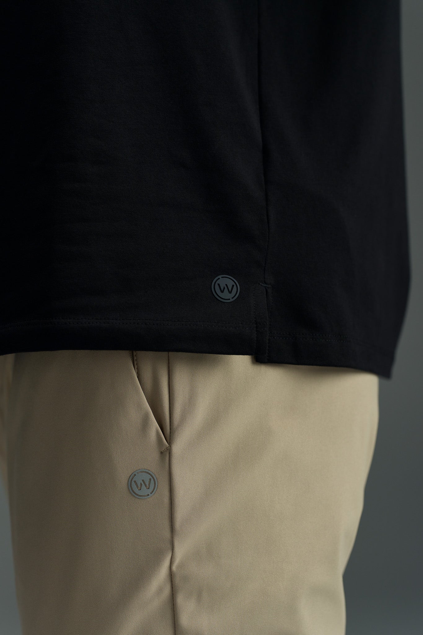A close-up of a person wearing the WearEver Outbound Polo in black and beige pants, both by WearEver, made from breathable cotton-blend fabric with small round logos near the hem and pocket, set against a plain gray background.