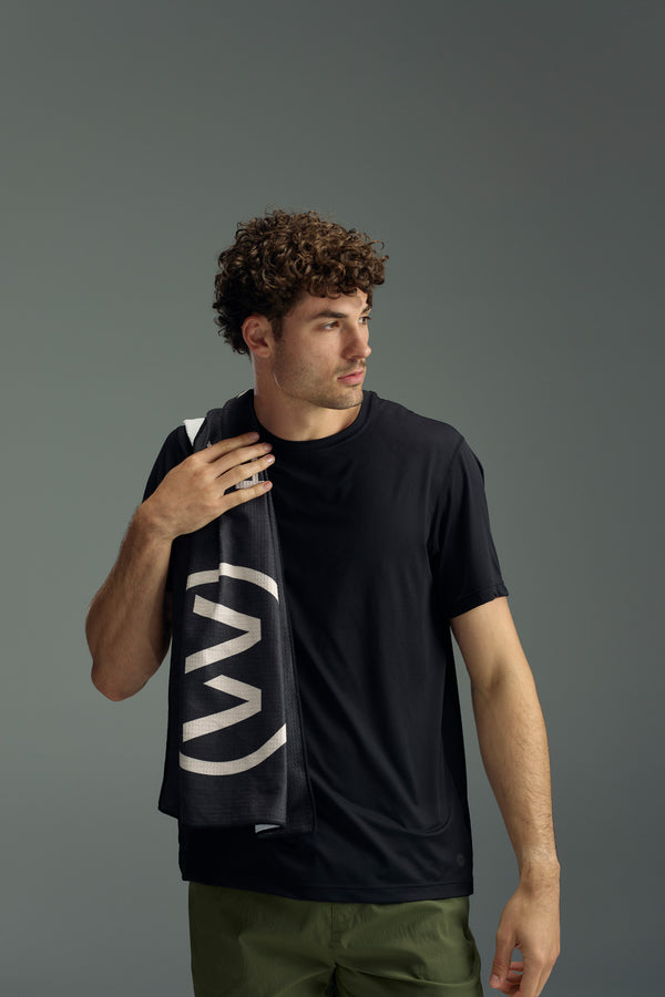 A young man with curly hair stands against a gray background, wearing a black t-shirt and green pants. He has the WearEver Utility Towel by WearEver, featuring white lettering, draped over his shoulder and gazes off to the side.