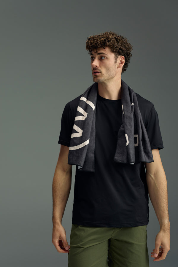 A man with curly hair, dressed in a black t-shirt and olive green pants, stands against a gray background with the WearEver Utility Towel from WearEver draped around his neck, looking to the side.