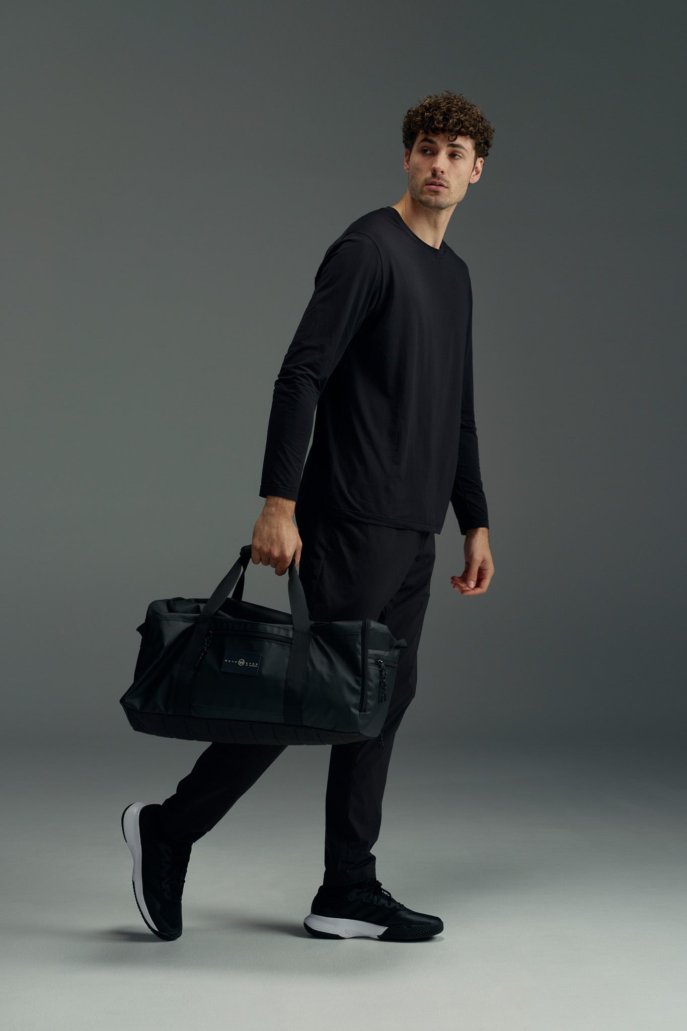 A man with curly brown hair, wearing a black long-sleeve shirt, black pants, and black sneakers, holds the WearEver Transit Holdall by WearEver in his left hand while looking right against a plain gray background.
