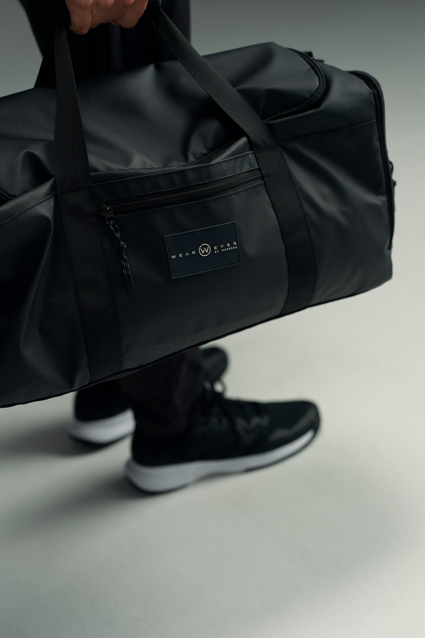 A person in black sneakers and dark pants holds the WearEver Transit Holdall, a sleek, water-resistant black duffel bag with front zipper pocket, sneaker pocket, and rectangular logo patch by WearEver, standing on a light gray surface.