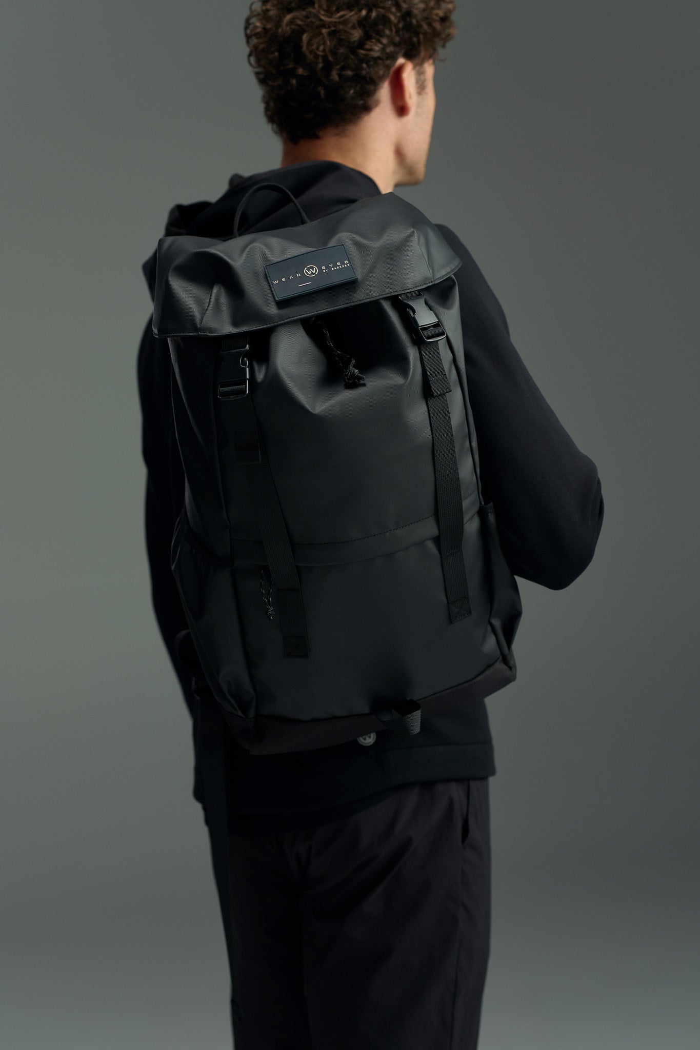 A person with curly brown hair in black attire is seen from behind wearing the WearEver Transit Backpack, which features a rectangular label and water-resistant zips, set against a neutral gray background.