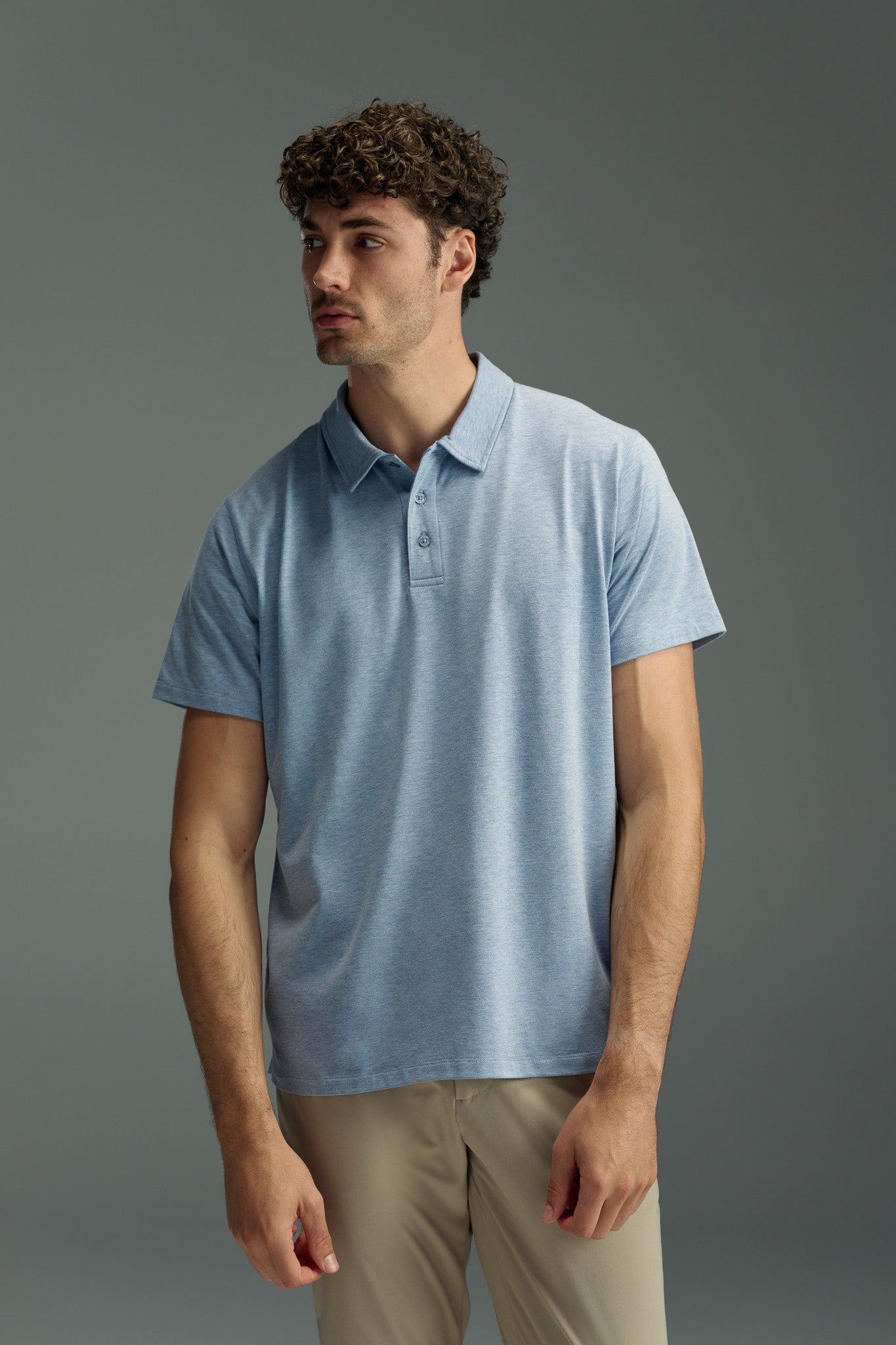 A man with curly brown hair wears a light blue WearEver Outbound Polo by WearEver, crafted from breathable cotton-blend fabric, and beige pants. He stands against a plain gray background, looking slightly to the side.