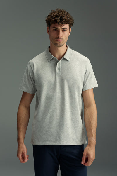 A man with curly brown hair wears the WearEver Outbound Polo by WearEver—a light gray, breathable, quick-drying, stretch fabric shirt—and dark pants, standing against a plain gray background, facing the camera with a neutral expression.