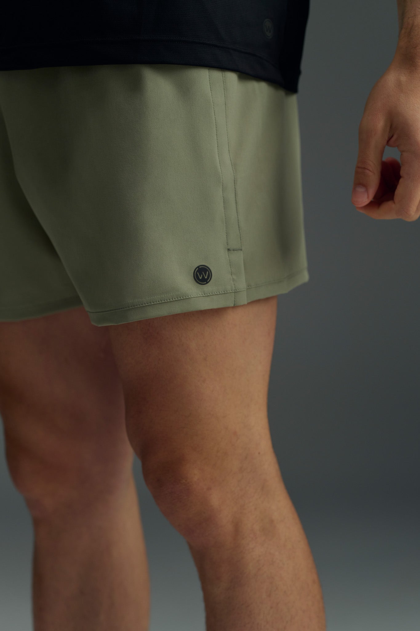 A person wears olive green WearEver Cadence Shorts (5" inseam) and a black top, shown waist to mid-thigh with one hand relaxed at their side against a plain background. The lightweight shorts appear breathable.