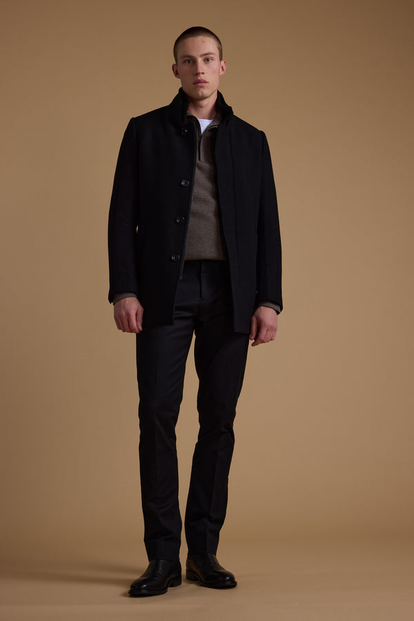A young man stands against a plain beige background, wearing the Barkers Raymond Coat—a black wool blend—over a brown sweater, white shirt, black pants, and black shoes. He faces forward with a neutral expression.