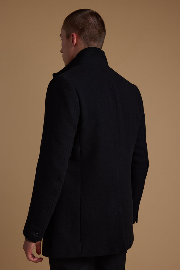 A person with short hair is shown from behind, wearing the Barkers Raymond Coat—a dark wool blend with a high collar—standing against a plain beige background.