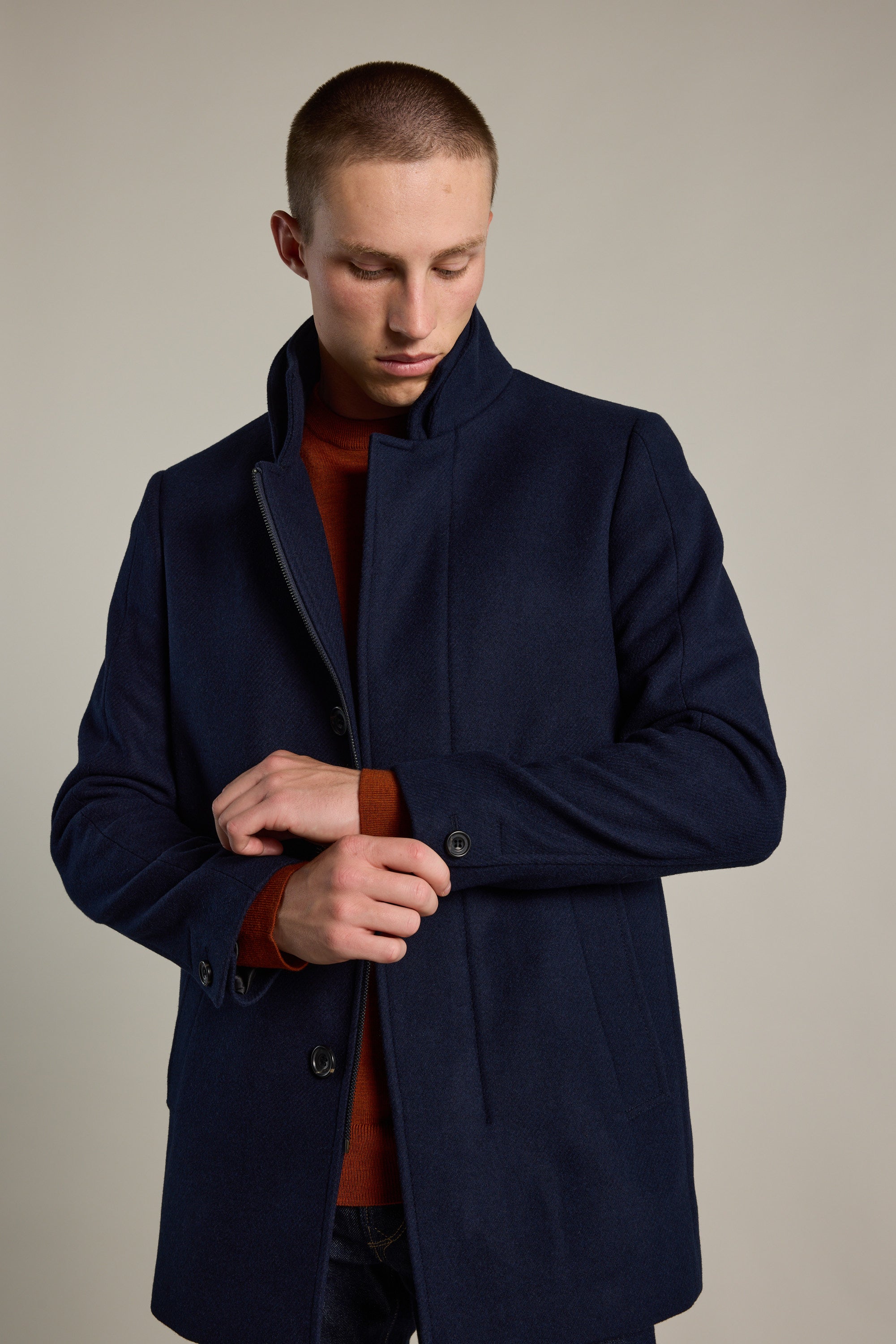 A young man with short hair wears the Barkers Raymond Coat, a dark navy wool blend, over a rust sweater. He looks down as he fastens the cuff of his coat against a plain, neutral background.