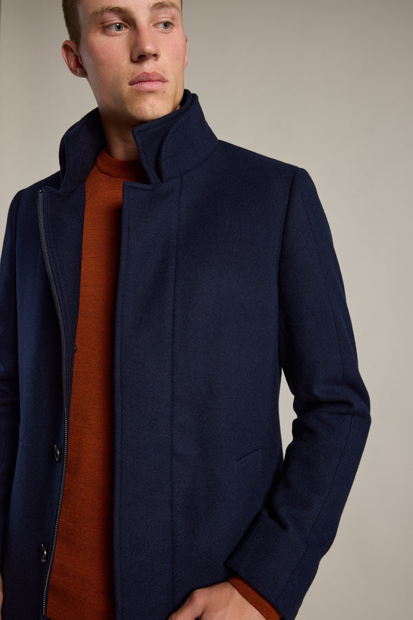 A person in the Barkers Raymond Coat, a dark blue men’s overcoat, is seen over an orange-brown sweater against a plain light background, gazing slightly to the side with a neutral expression.