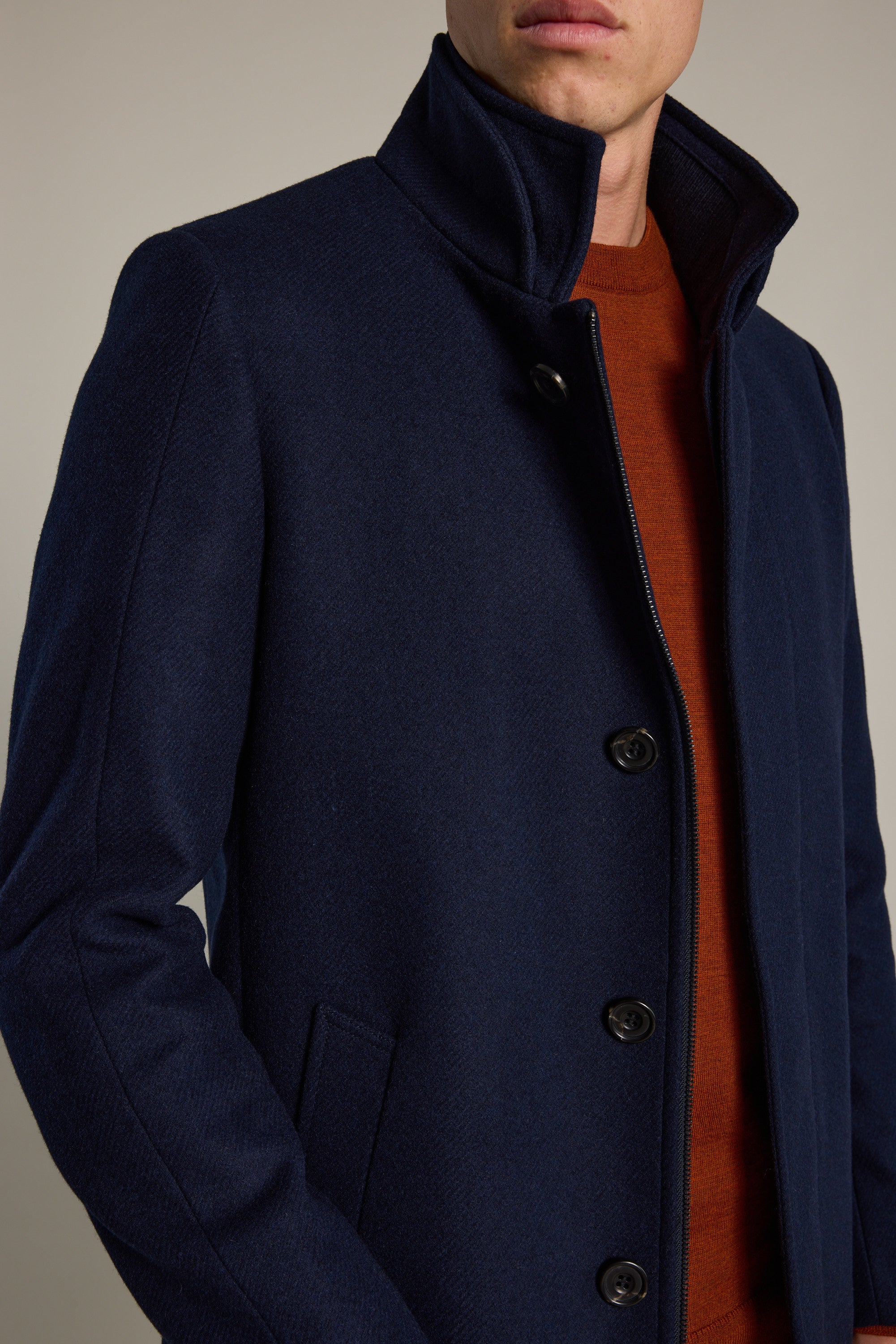 A person wears the Raymond Coat by Barkers, a dark navy wool blend with a high collar and black buttons, layered over an orange sweater, photographed from shoulders to waist against a plain background.