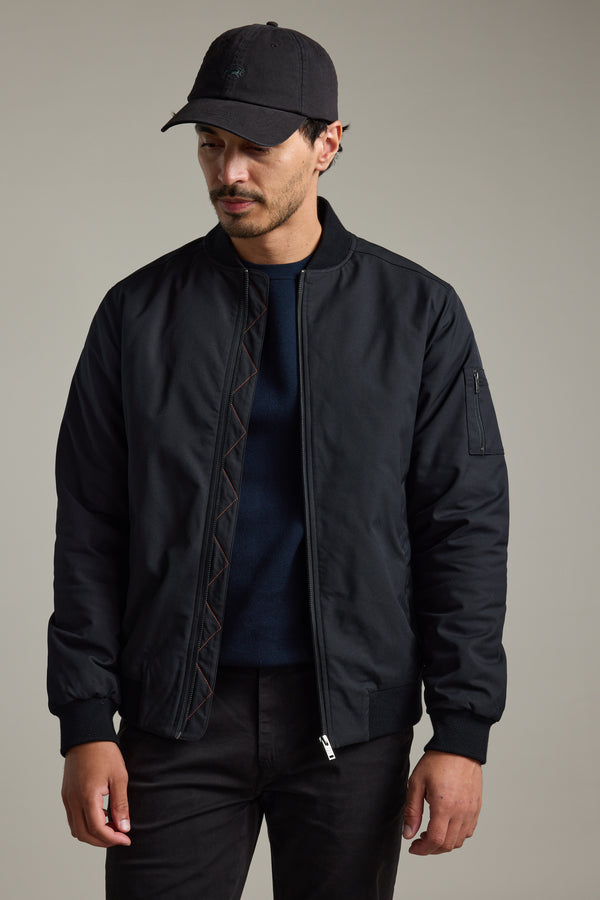 Man in a Barkers Dunhill Bomber Jacket, black pants, and cap stands against a plain background.
