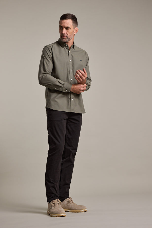 A man stands against a plain background wearing an olive green button-up, Barkers Carnaby Chino trousers made from responsibly sourced cotton, and light brown shoes, looking slightly left with one hand adjusting his cuff.
