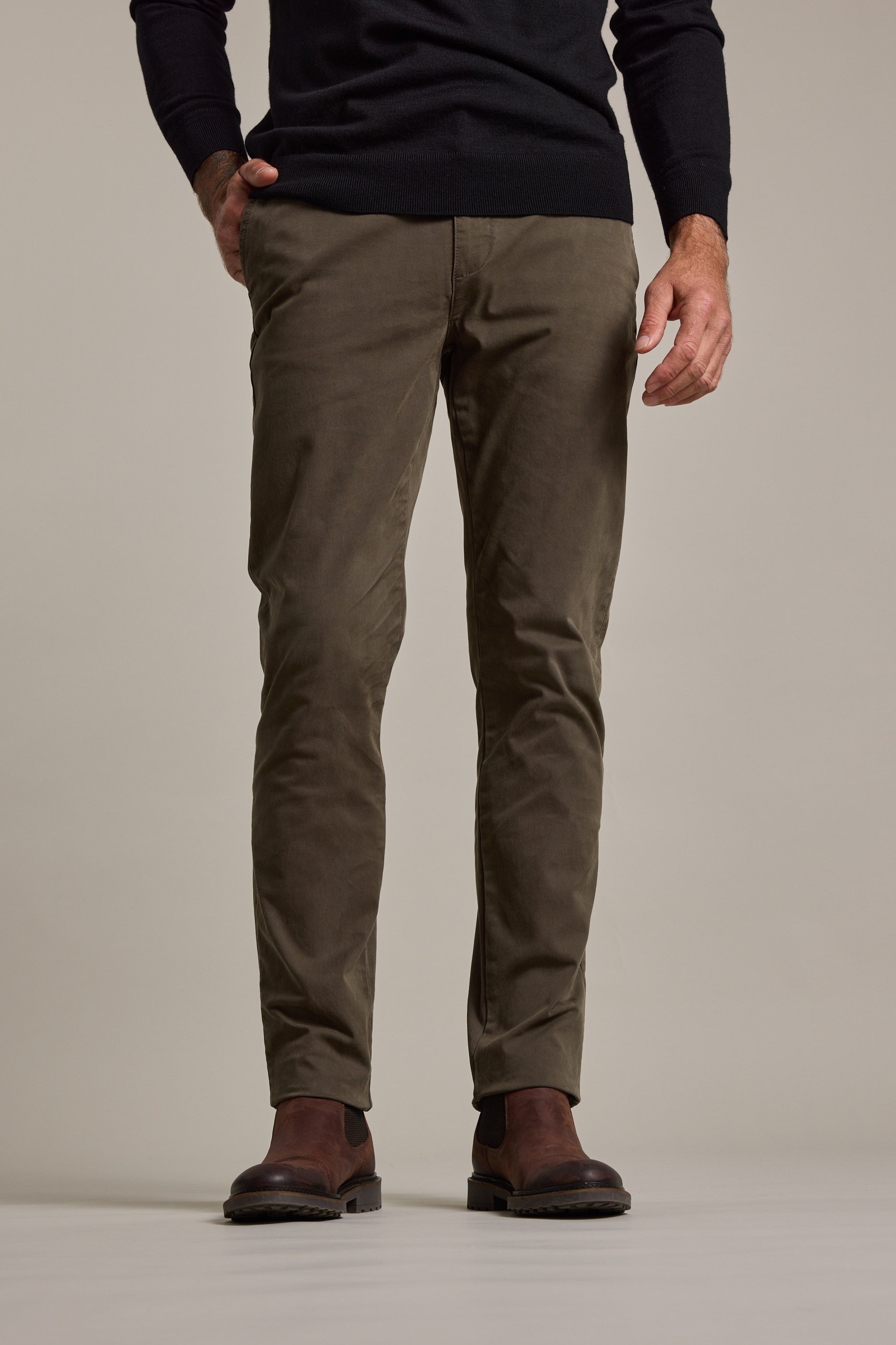 A person wears Barkers' Carnaby Chino in olive green, made from responsibly sourced cotton, styled with a black sweater and brown leather shoes. Only their lower body is visible against a plain light background.