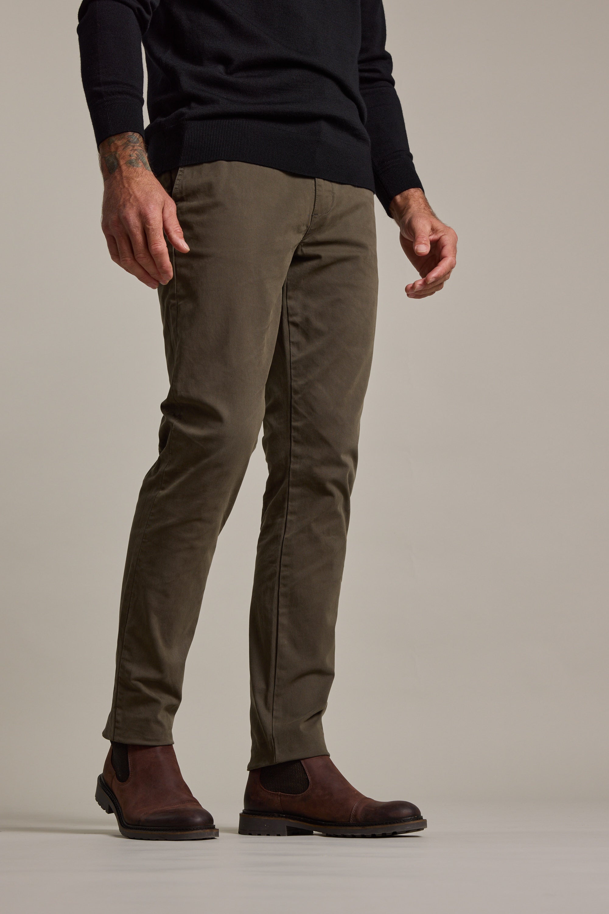 A person stands against a neutral background, wearing Barkers Carnaby Chino in olive green, a black long-sleeve sweater, and brown Chelsea boots. The photo is cropped from mid-torso down.