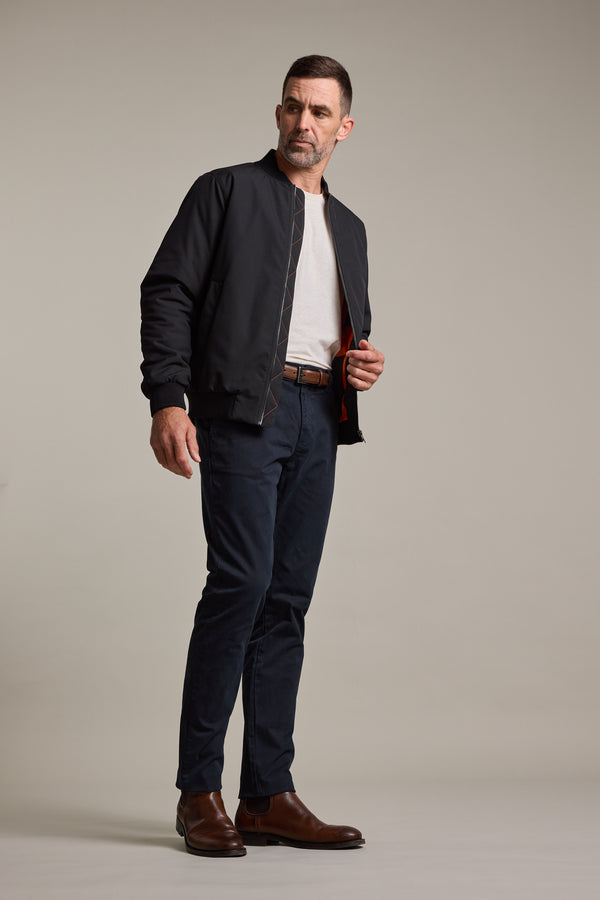 A man poses against a plain light gray background in Barkers Carnaby Chino trousers, a black jacket, white t-shirt, and brown leather shoes, looking slightly left while adjusting his jacket.