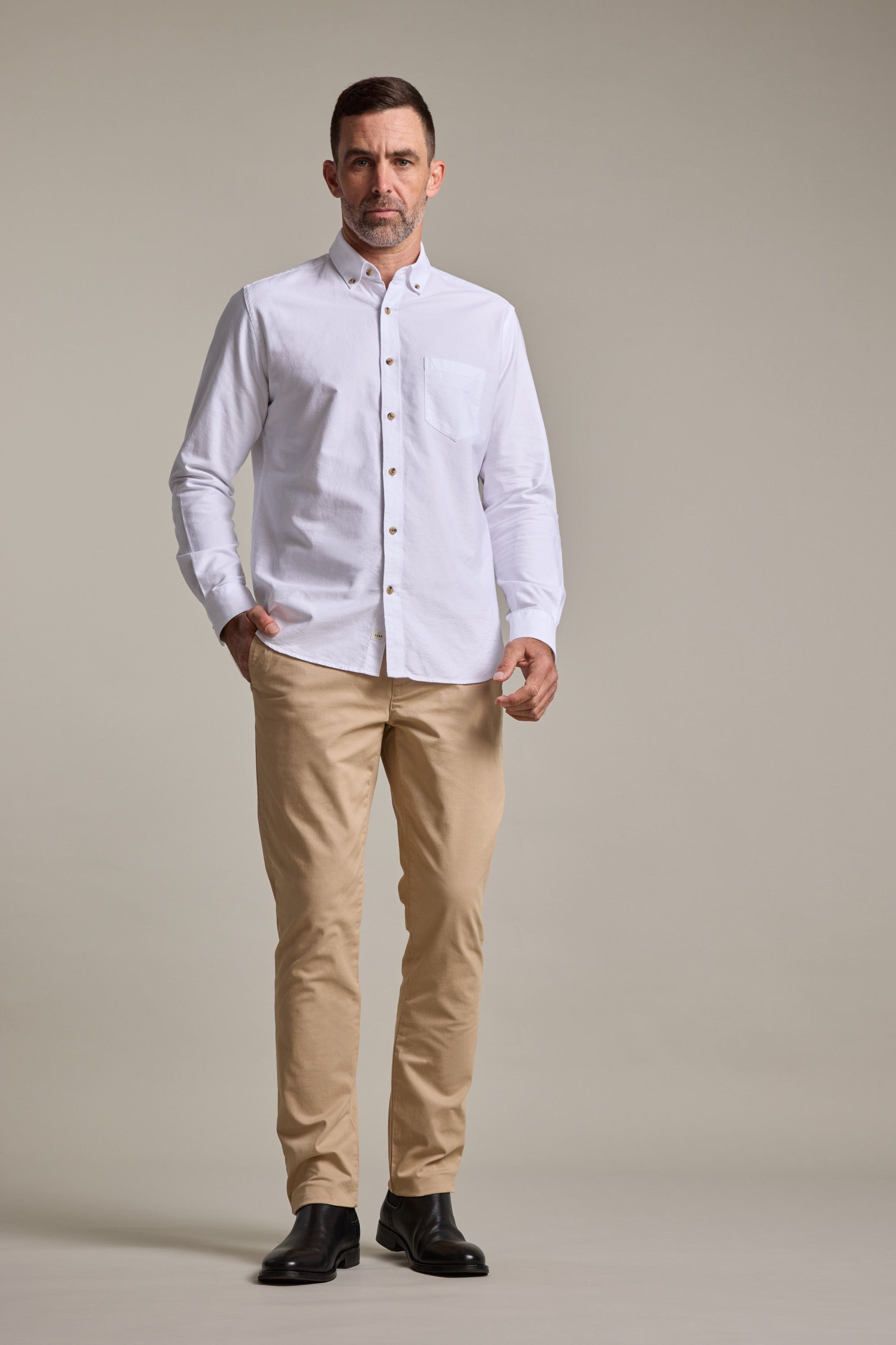 A man stands facing forward wearing a white button-up shirt, Barkers Carnaby Chino beige pants made from responsibly sourced cotton, and black shoes against a plain light background.