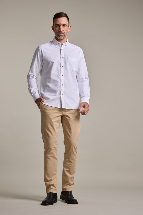 A man stands facing forward wearing a white button-up shirt, Barkers Carnaby Chino beige pants made from responsibly sourced cotton, and black shoes against a plain light background.