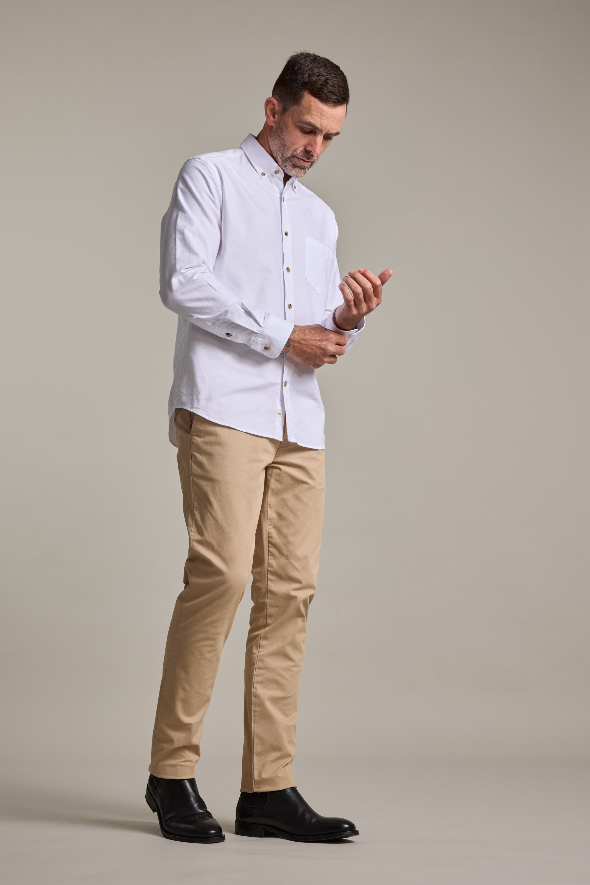A man stands against a plain background, looking down as he buttons the cuff of his white dress shirt. He is wearing Barkers Carnaby Chino pants made from responsibly sourced cotton and black shoes.