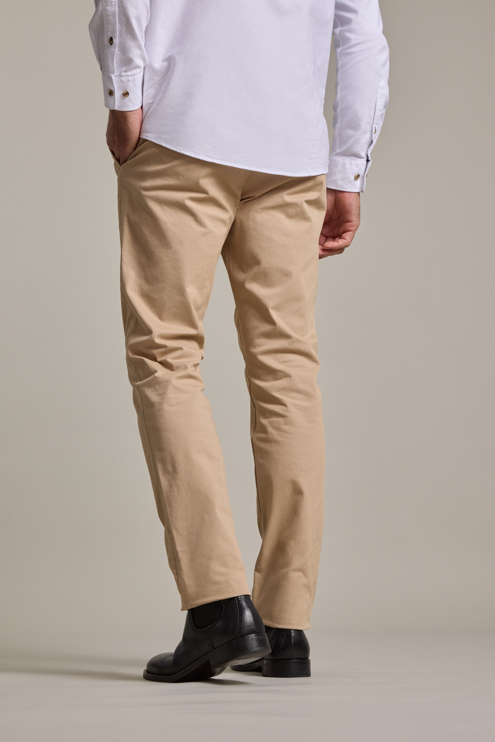 A person stands with their back to the camera, wearing a white button-up shirt and Barkers Carnaby Chino men's stretch chinos made from responsibly sourced cotton, paired with black shoes against a plain background.