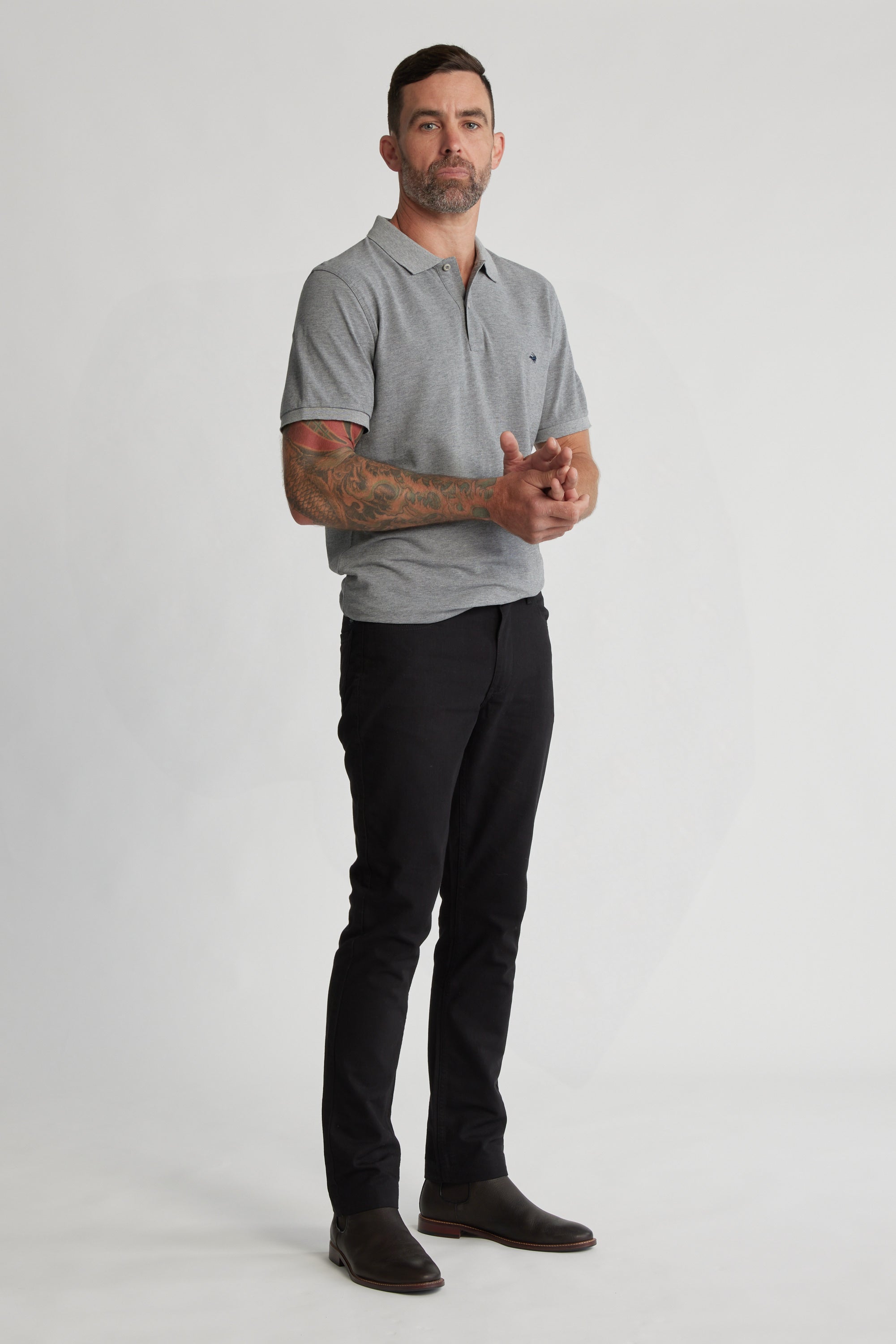 A man with short dark hair and a beard stands against a plain background, wearing a gray polo shirt, Barkers Oxmor 5 Pkt Pant O in black, dark shoes, and showing tattoos on his left forearm while holding his hands together.