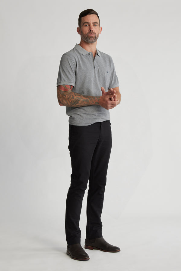 A man with short dark hair and a beard stands against a plain background, wearing a gray polo shirt, Barkers Oxmor 5 Pkt Pant O in black, dark shoes, and showing tattoos on his left forearm while holding his hands together.