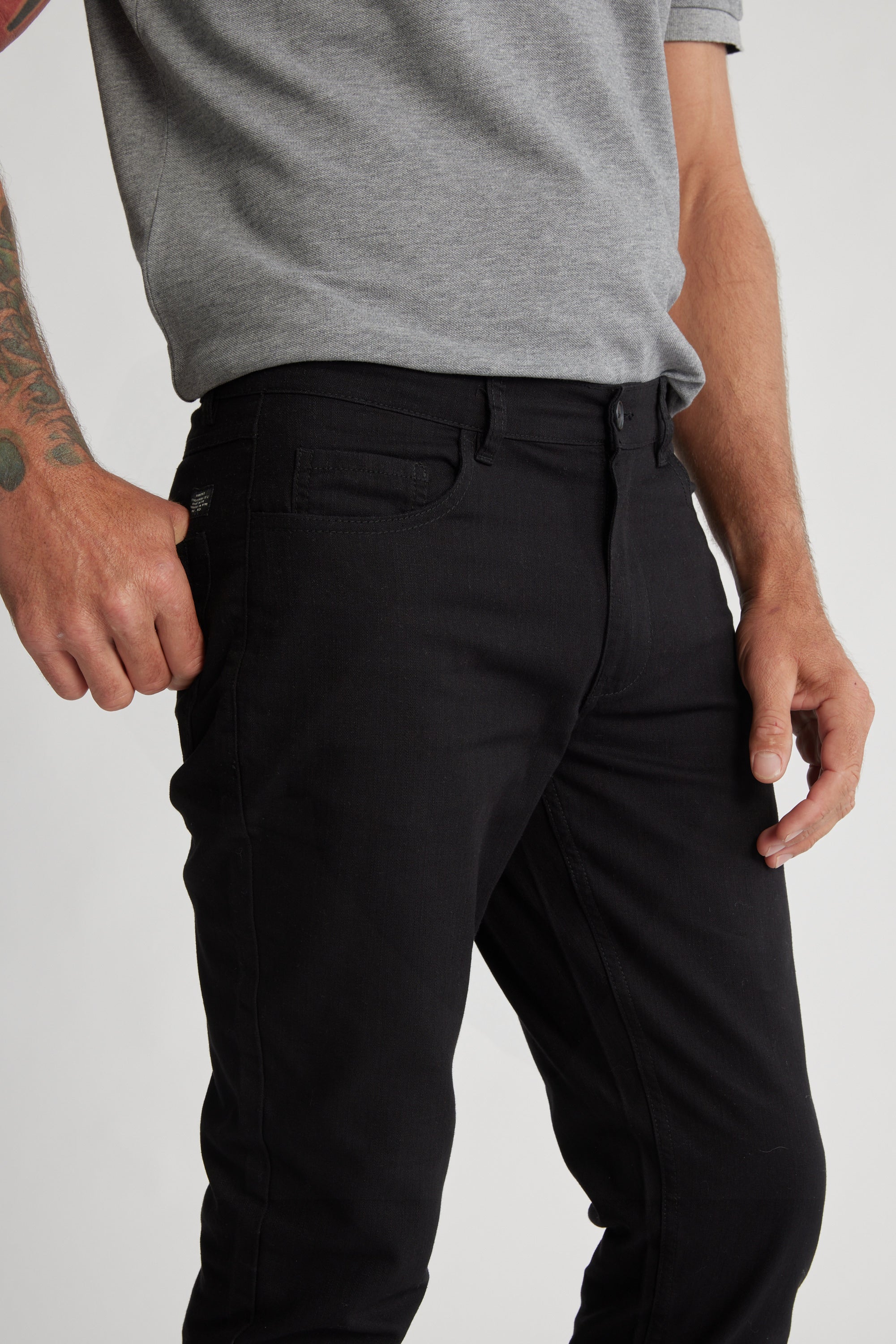 A person in a grey short-sleeve shirt and Barkers Oxmor 5 Pkt Pant O (black, taper fit) stands with a tattooed arm near their pocket. Image shows from shoulders to mid-thigh.