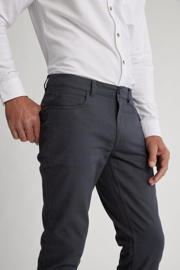 A person wears Barkers' Oxmor 5 Pkt Pant O in dark gray with a white button-up shirt, standing with one hand in a pocket and the other by their side against a plain light background.