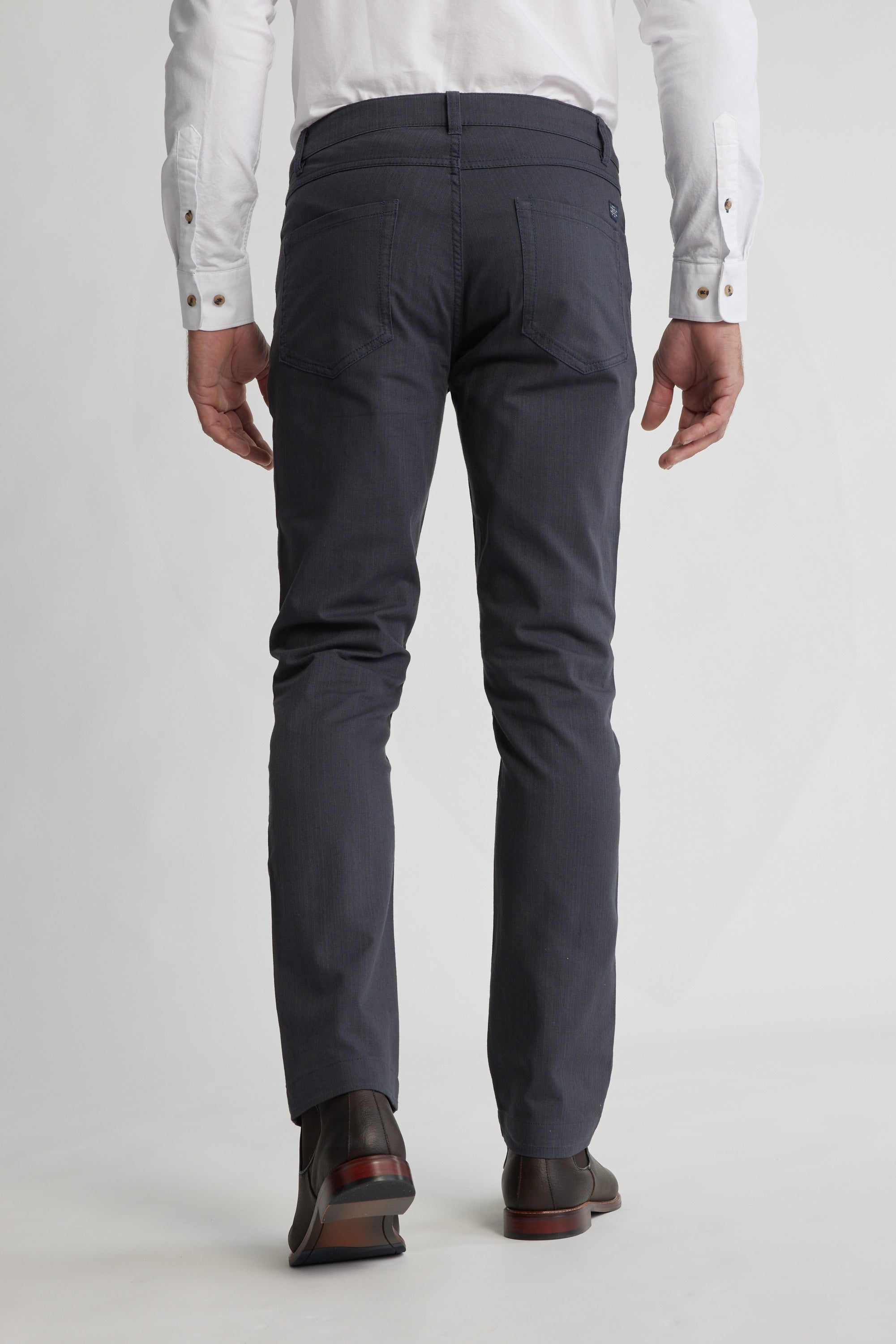 A person wearing a white dress shirt and Barkers Oxmor 5 Pkt Pant O in dark gray with black dress shoes is shown from the back, standing against a plain light-colored background to highlight the pants’ rear view.