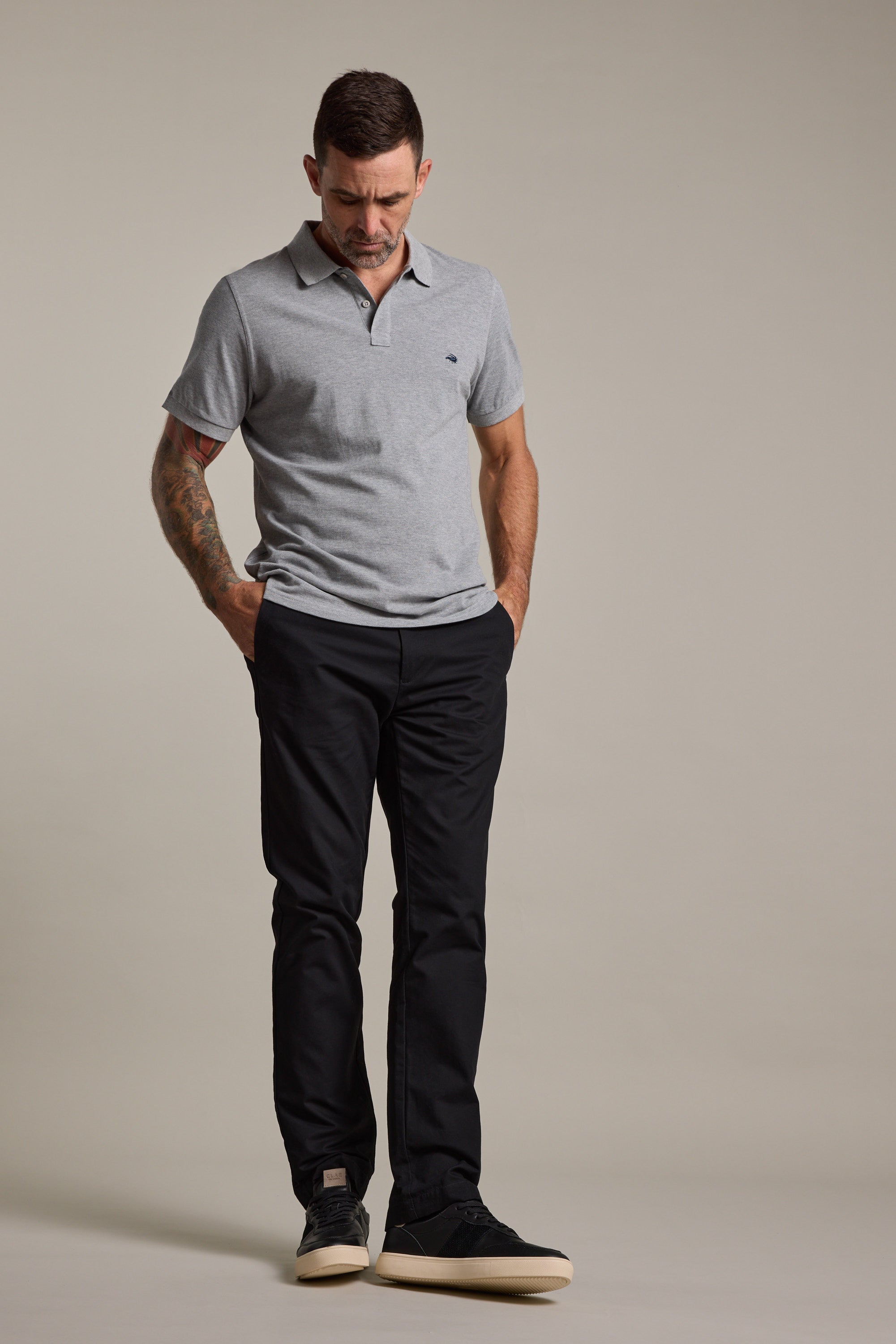 A man stands with his hands in his pockets, looking down. He wears a light gray polo, Barkers Dundas Chino trousers in a relaxed fit, and black shoes against a plain neutral background.