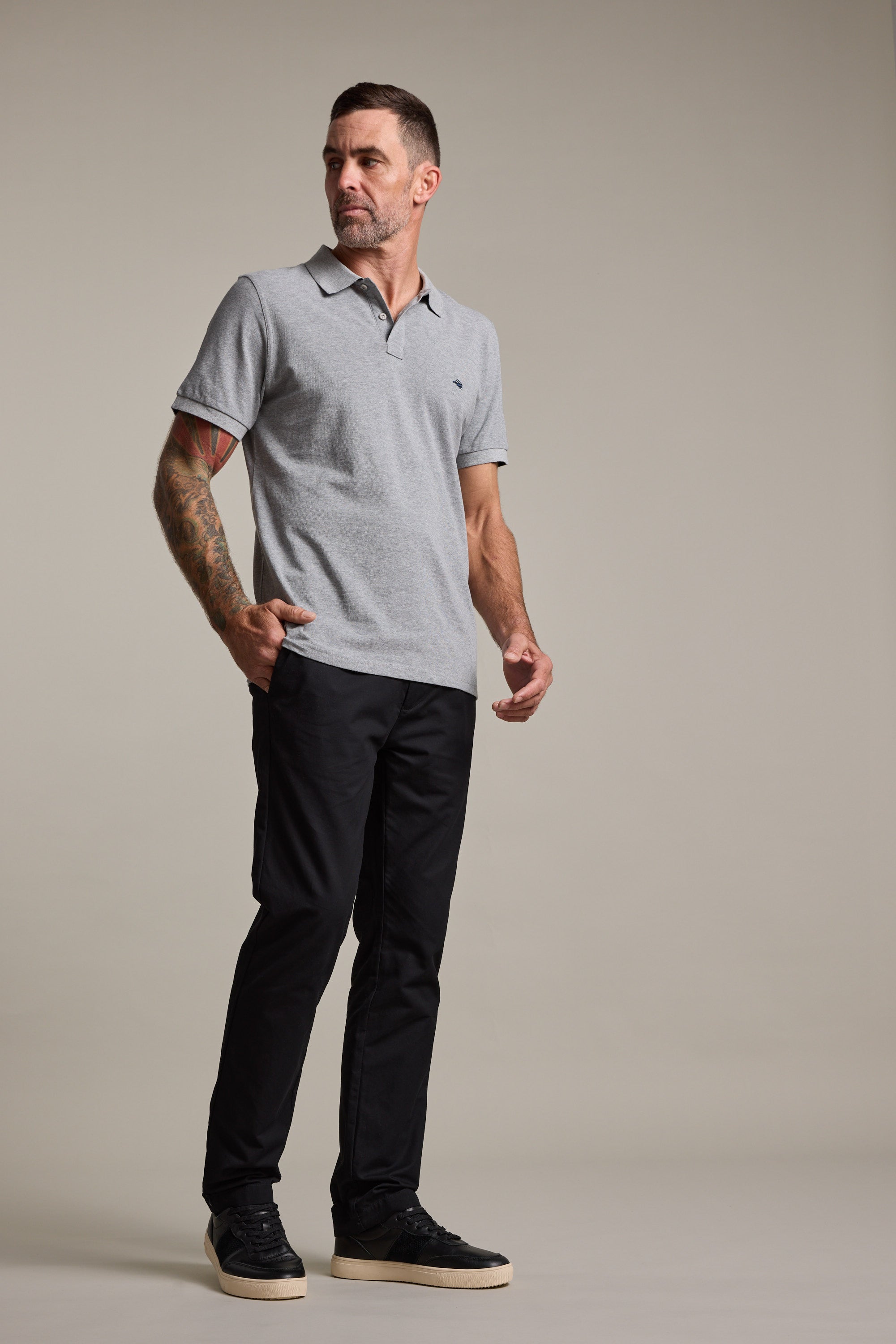 A man with short hair and tattoos on his left arm stands against a plain background, wearing Barkers Dundas Chino trousers with a gray polo shirt and black sneakers with white soles. He looks to his left, expression neutral.