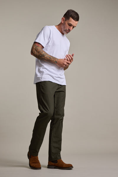 A tattooed man wears a relaxed fit white t-shirt, Barkers Dundas Chino olive green cotton twill trousers, and brown boots. He stands against a plain background, looking down with his hands together.