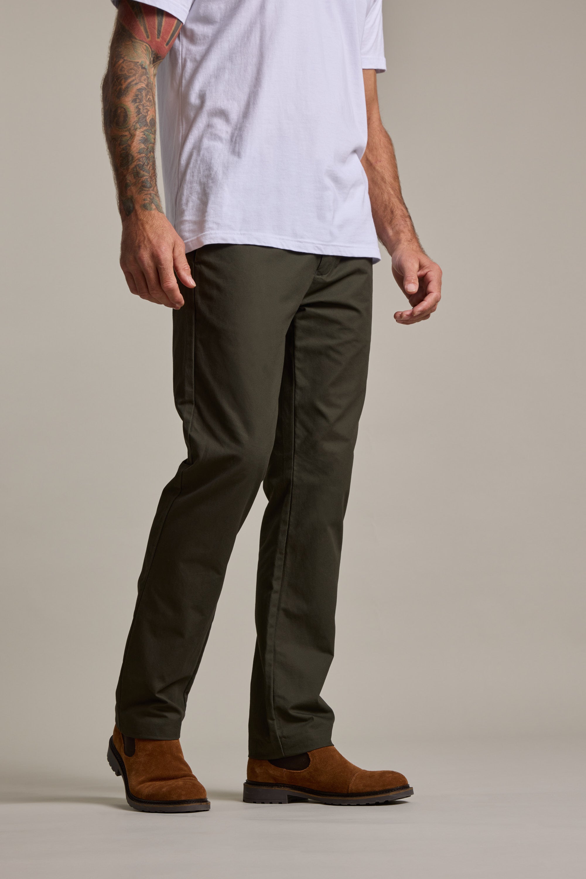 A person stands against a plain background, showing only the lower half in a white t-shirt, Barkers Dundas Chino trousers in olive green, and brown suede boots. One visible arm is decorated with a colorful tattoo.