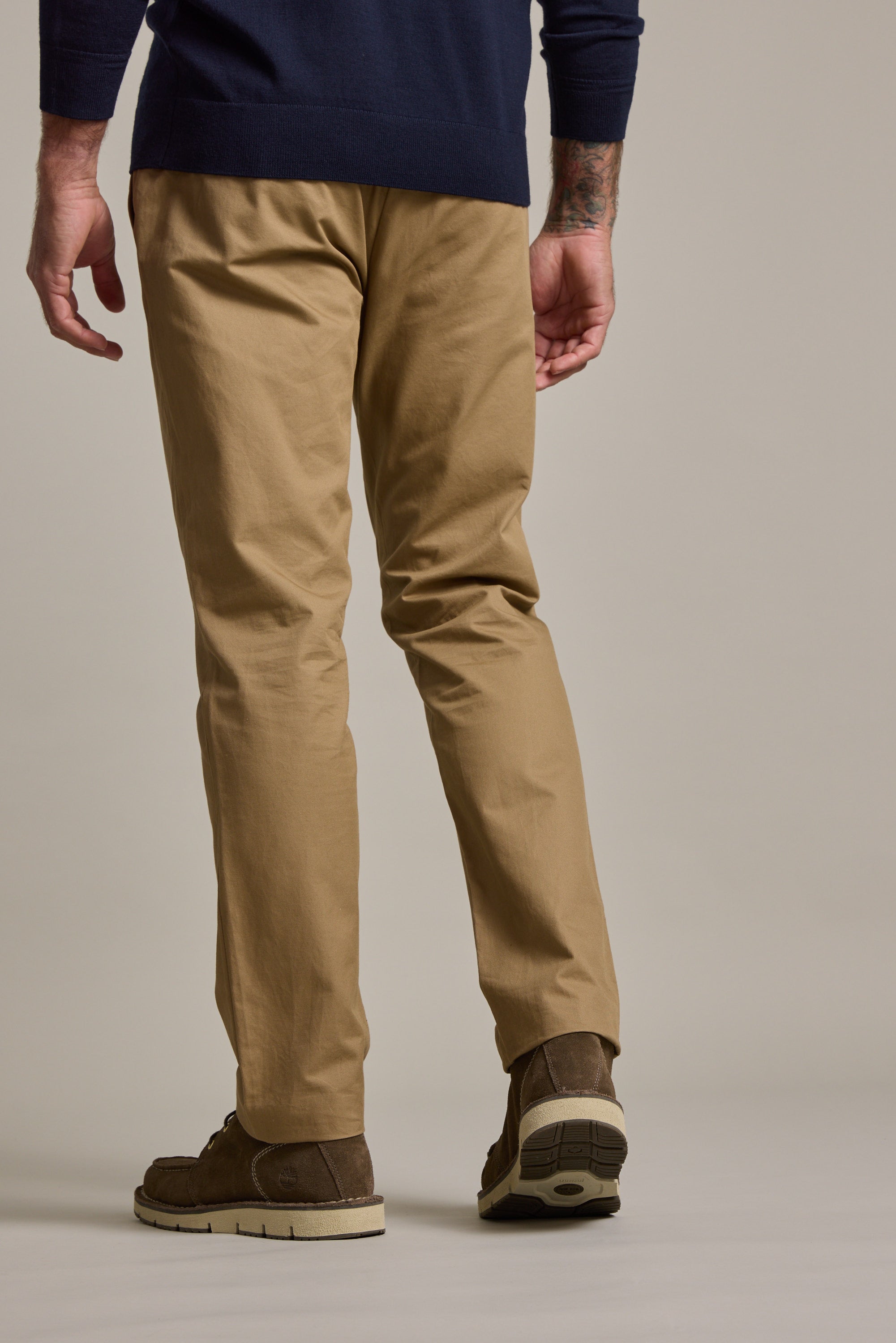 A person is seen from the back, wearing Barkers Dundas Chino trousers in lightweight cotton twill, brown shoes, and a navy sweater, standing against a plain background.