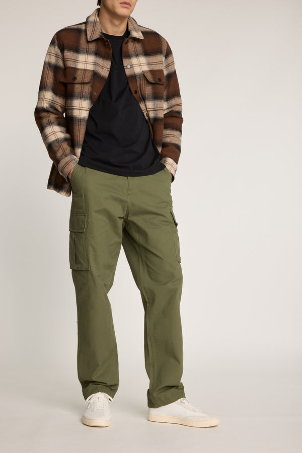 A man stands against a plain background with his hands in his pockets, wearing a brown plaid overshirt, black t-shirt, Barkers Barnard Cargo Pant in a relaxed fit, and white sneakers. His face is not fully visible.
