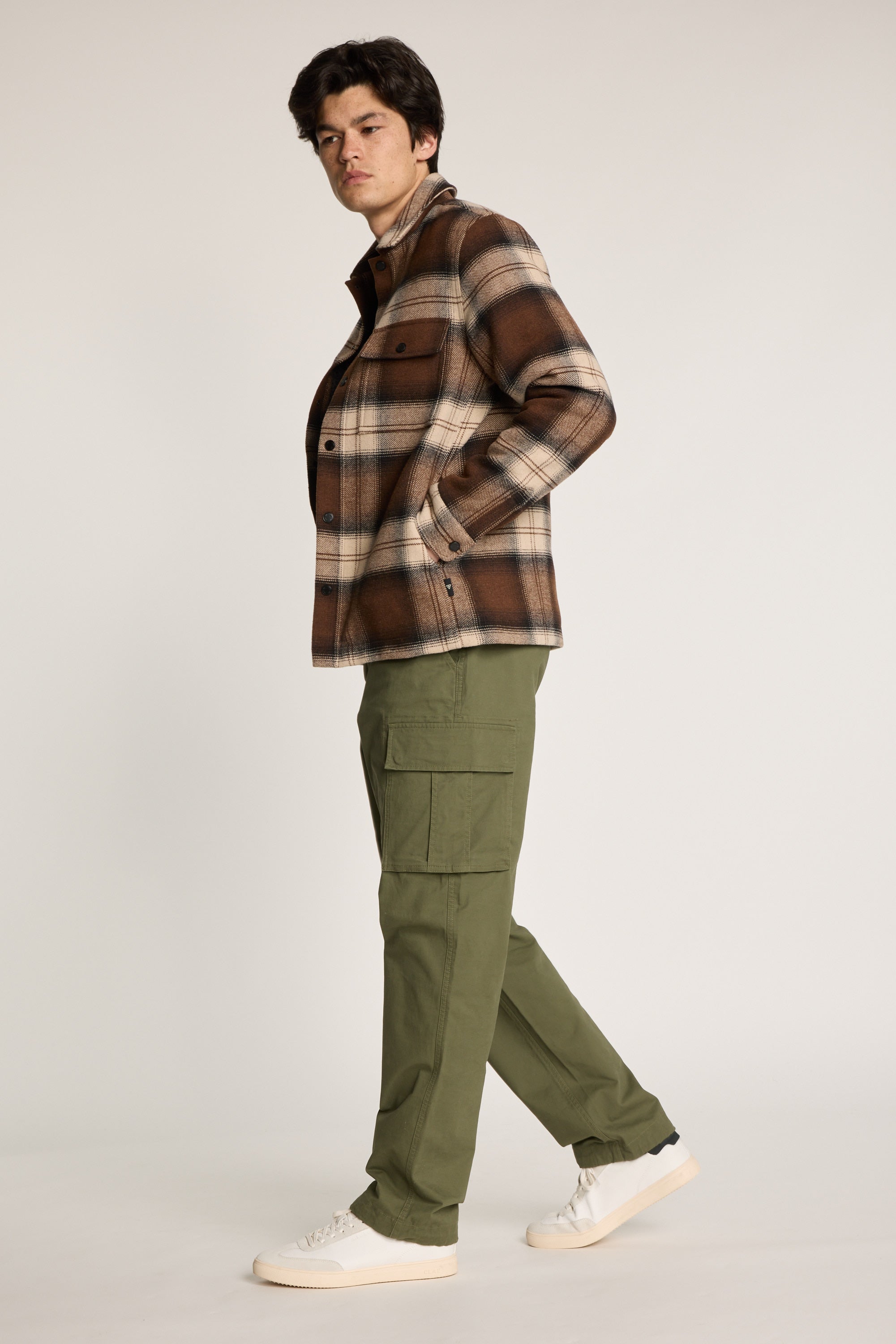 A man in Barkers Barnard Cargo Pant, a brown plaid jacket, and white sneakers stands against a plain light background, looking sideways while taking a step.