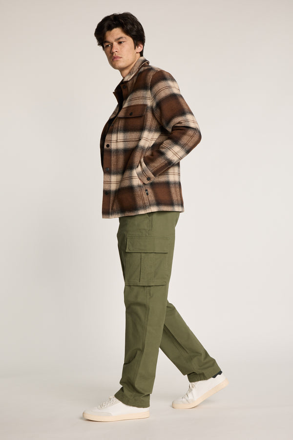 A man in Barkers Barnard Cargo Pant, a brown plaid jacket, and white sneakers stands against a plain light background, looking sideways while taking a step.