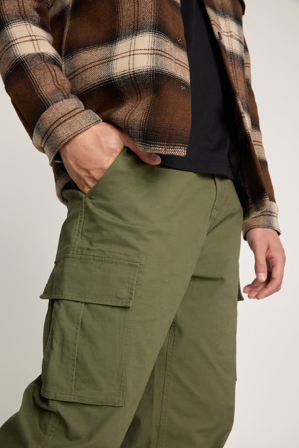 A person in Barkers Barnard Cargo Pant, relaxed fit olive green twill cotton pants, paired with a brown and beige plaid shirt over a black top, shown from shoulders to thighs with one hand in the pocket.