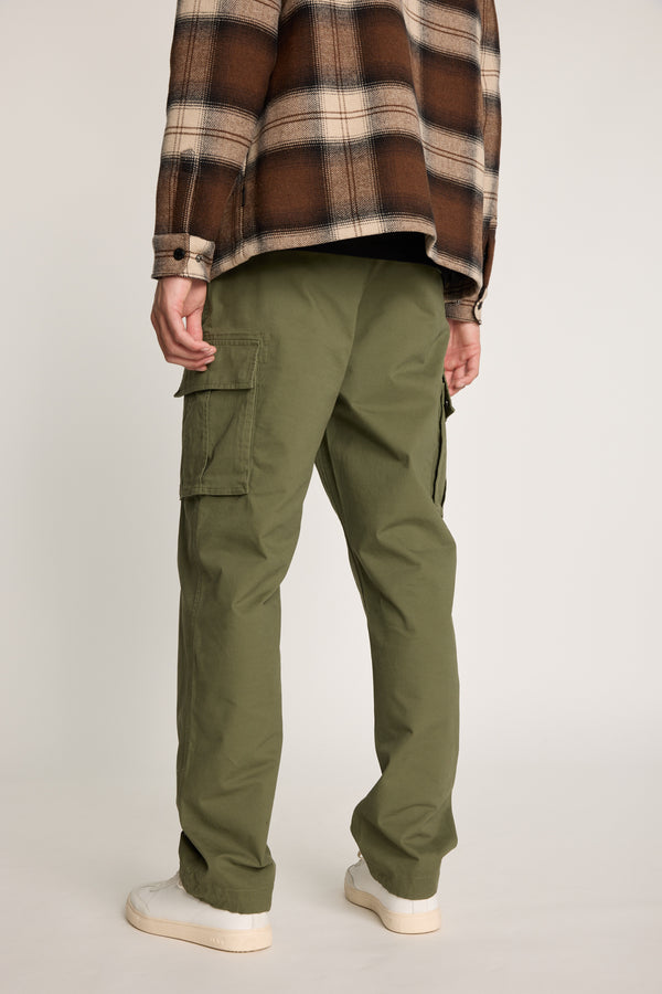 A person wears Barkers Barnard Cargo Pant in olive green twill cotton, paired with a brown plaid shirt and white sneakers, standing facing away with hands at their sides against a plain background.