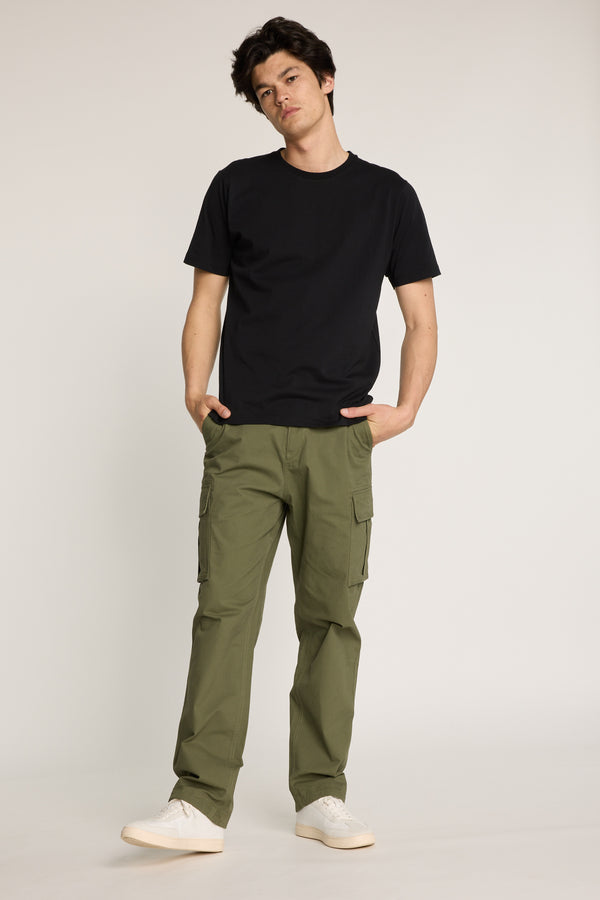 A person stands against a plain background wearing the Barkers Barnard Cargo Pant in relaxed fit olive green twill cotton, paired with a black t-shirt and white sneakers, hands in pockets and a neutral expression.