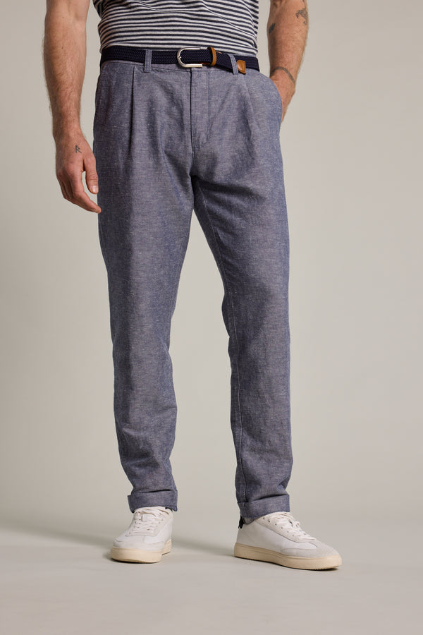 A man stands against a plain light background, hands in pockets, wearing Barkers' Newman Linen Pants with a striped shirt tucked in, black belt, and white sneakers.