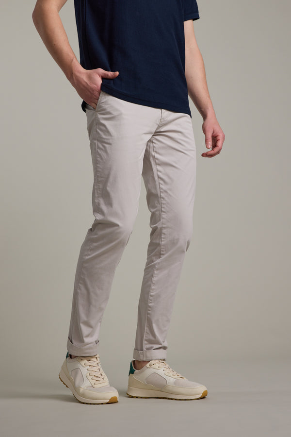 A person, shown from the waist down, stands against a plain light background wearing Barkers Kingly Chino Pant in light beige, paired with a navy blue t-shirt and cream sneakers; one hand is in their pocket.